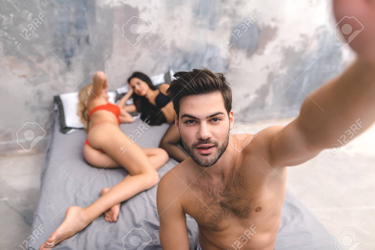 Man Wake Up After Night Threesome And Take Selfie. Two Girls Sleeping In Bedroom While Man Wake Up And Make Selfie. Sex Party Concept. Stock Photo Picture and Royalty Free Image. Image