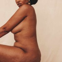 Confident young woman sitting naked in a studio. Body positive young woman sitting sideways against a studio background. Young woman feeling comfortable in her own body and curves. stock photo